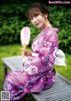 A woman in a purple kimono sitting on a bench holding a fan.